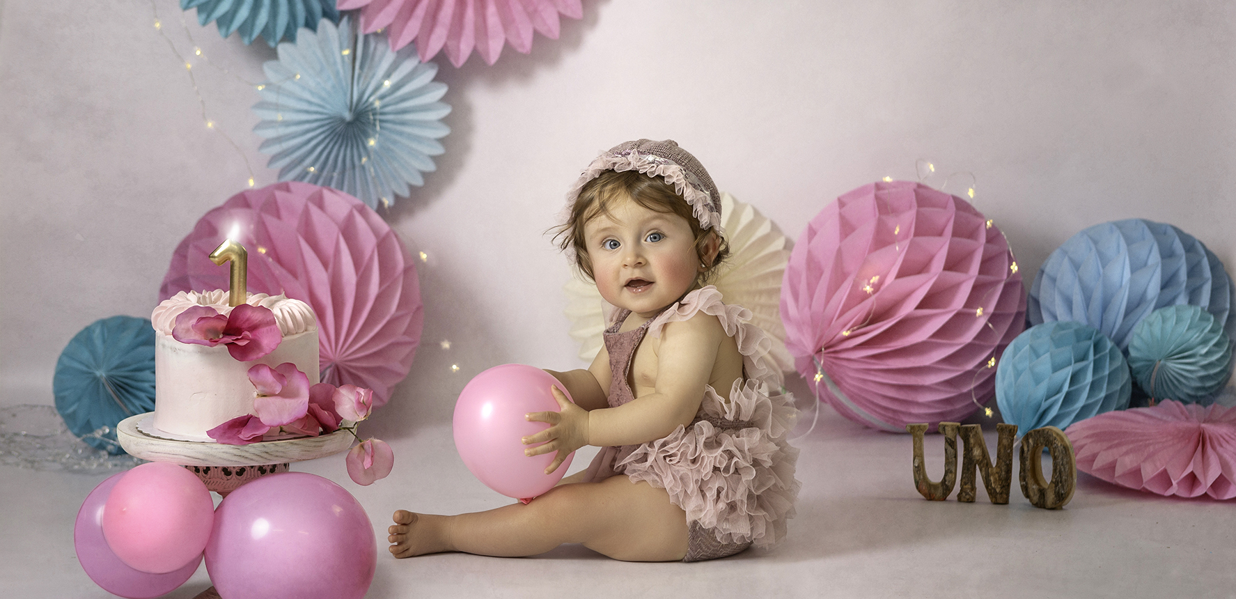Fotografía de una niña con vestido rosa con una tarta con un uno sujetando un globo mirando a la cámara